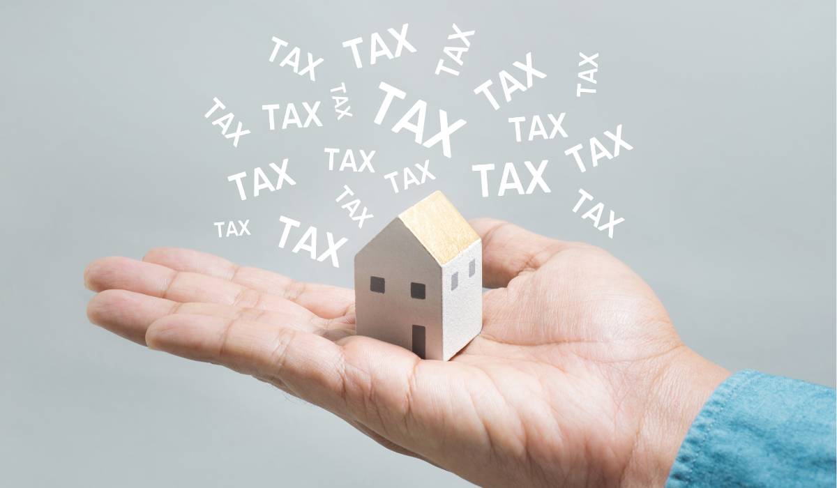 A hand holding a miniature home with multiple "tax" words coming out of it on a grey background